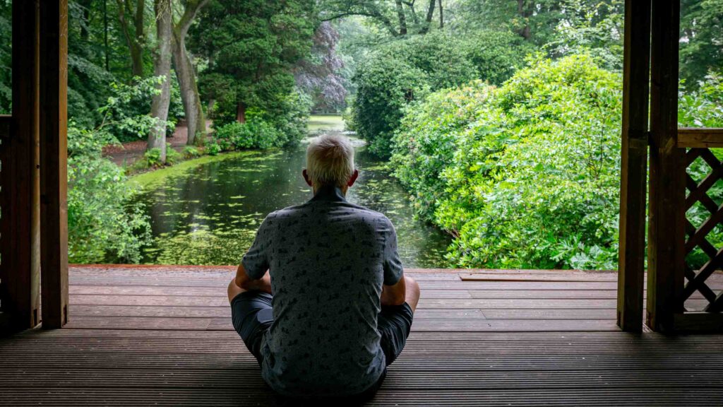 Man sitting on wooden deck meditating with pond and trees front of him